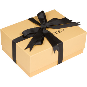 A closed, rectangular beige gift box features branded packaging with a glossy black ribbon tied into a bow on top. The box has subtle text partially visible under the ribbon.