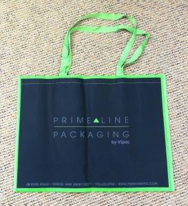 A blue and green tote bag with Prime Line Packaging by Vipac printed in the center, perfect for a trade show. The bag has two sturdy handles and rests on a textured beige carpet. Contact information is neatly printed along the bottom edge.