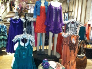 A boutique display features mannequins dressed in vibrant dresses of purple, orange, and blue. Nearby racks and hangers showcase an array of dresses along with a denim jacket. A rustic wooden floor and staircase add charm to the background.