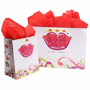 Two gift bags with colorful designs labeled Sassy Designs filled with red tissue paper, adorned with branded ribbons. The larger bag is behind the smaller one, both featuring a vibrant and artistic pattern with a playful aesthetic.