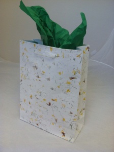 A decorative gift bag crafted from hand-made papers, featuring a floral pattern and green tissue paper peeking out from the top, placed against a plain background.