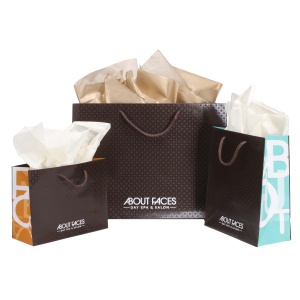 Three gift bags labeled About Faces Day Spa & Salon showcase their elegant design services. Filled with white and beige tissue paper, the largest bag claims the spotlight at the center, flanked by two smaller ones on each side.