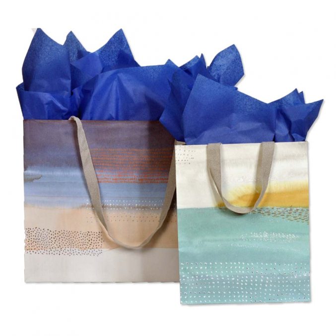 Two decorative gift bags with blue tissue paper. The larger bag has a gradient of brown and blue colors, while the smaller bag features green and yellow hues. Both have beige handles and subtle dot patterns.