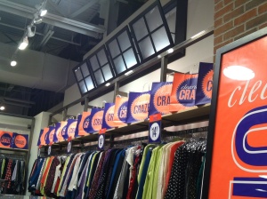 A clothing store with colorful shirts and tops arranged on racks below signage featuring repeated CRA text in orange and blue. The ceiling boasts industrial-style lighting, complemented by sleek in-store displays positioned above the shelves for a modern shopping experience.