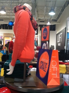 A chic in-store display features a mannequin dressed in an orange sweater and plaid scarf, capturing the essence of LOFT CRAZE. Behind it, neatly folded clothes fill the shelves, inviting customers to explore further.