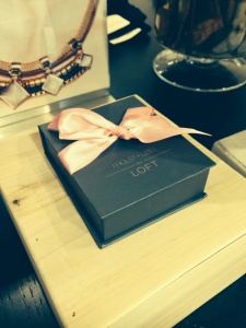 A small, closed dark gift box wrapped with a pink ribbon is displayed on a wooden surface, symbolizing support for Breast Cancer Awareness. Behind it, there's a blurred image of jewelry.