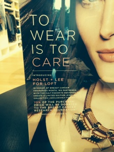 Close-up of a store advertisement featuring a partially visible woman wearing a chunky gold necklace. Text on the ad reads TO WEAR IS TO CARE and highlights a special collaboration for Breast Cancer Awareness, with 70% of proceeds supporting a research foundation.