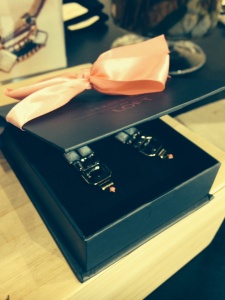A stylish black box with a pink bow, representing Breast Cancer Awareness, contains two neatly arranged cufflinks on a black velvet interior, displayed on a wooden surface.