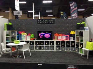 A trade show booth with a PrimeLine Packaging sign showcases colorful gift bags on white shelving units, reminiscent of the vibrant displays from trade shows 2015. A round table with chairs complements the setup, while a TV screen displays a logo against dotted-patterned walls and carpeted flooring.