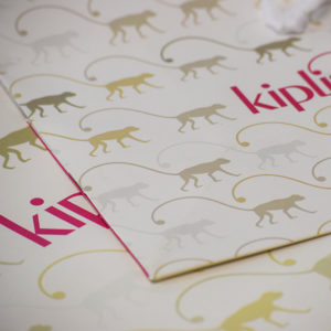 A close-up view of the paper reveals illustrations of monkeys in various shades of beige and gray. The word "kipling" appears partially in bold pink text, adding a lively contrast to the overall design.