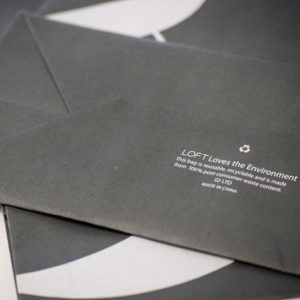 A close-up of a black paper bag with white text reading, LOFT Loves the Environment. This eco-friendly paper bag is reusable and recyclable, crafted from 100% post-consumer waste. Made in China, it proudly displays a recycling symbol above the text.