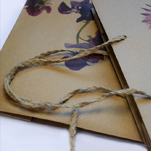 A close-up of a cardboard bag with a twisted jute handle and floral designs in purple and pink hues. The focus is on the textured handle, while the decorative patterns add a touch of color to the background.
