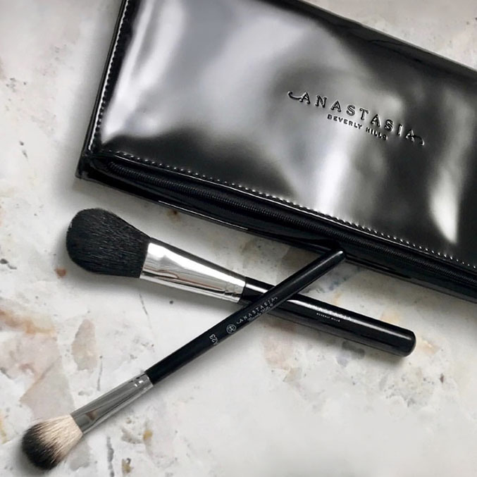 A shiny black Anastasia Beverly Hills makeup brush case with two makeup brushes placed diagonally across it. One brush has a fluffy black head, and the other has a white angled bristle head. They are on a marbled surface.