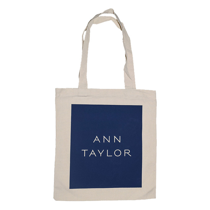 A beige tote bag with handles featuring a navy blue square on the front. Inside the square, the text ANN TAYLOR is printed in white, uppercase letters.
