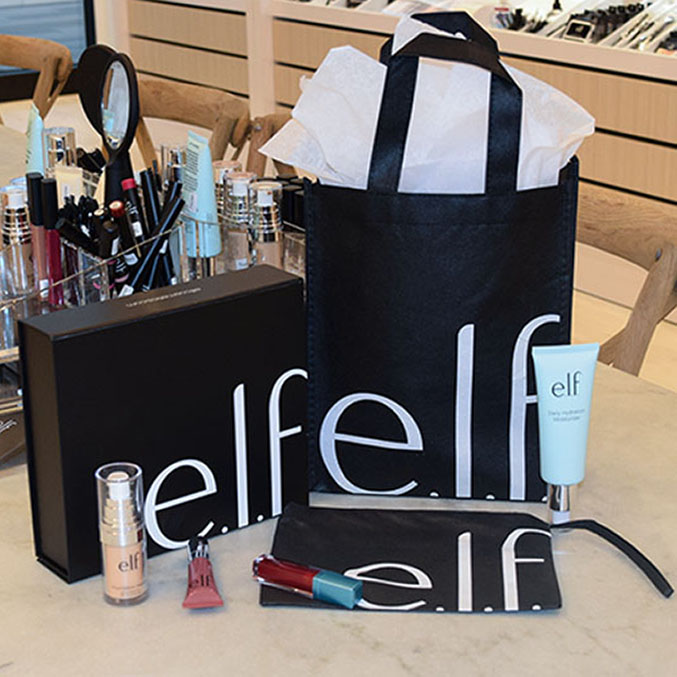 A display of e.l.f. cosmetics products on a wooden table. Items include a black shopping bag with white lettering, a foundation, lip gloss, pouch, and skincare tube. Mirrors and more products are seen in the background.