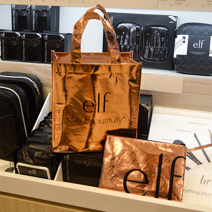 A display of e.l.f. cosmetics products, featuring rose gold bags with the e.l.f. play beautifully logo. The background shows various cosmetic kits and brushes in black packaging on shelves.