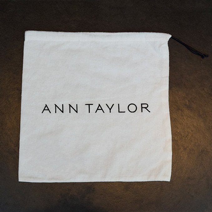 A white drawstring pouch with the text ANN TAYLOR in black, centered on the front, placed against a dark background.