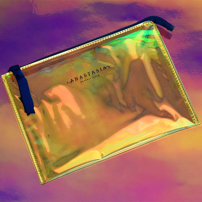 A holographic makeup bag with a black zipper and blue pull tab, labeled ANASTASIA BEVERLY HILLS, is placed on a colorful iridescent background.