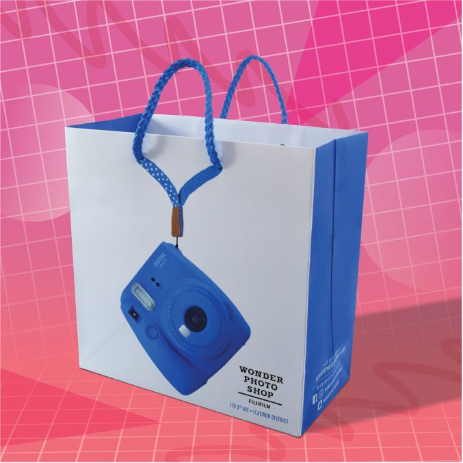 A shopping bag with a blue camera design and braided blue handles against a pink grid background. The bag reads Wonder Photo Shop and showcases a modern, vibrant style.