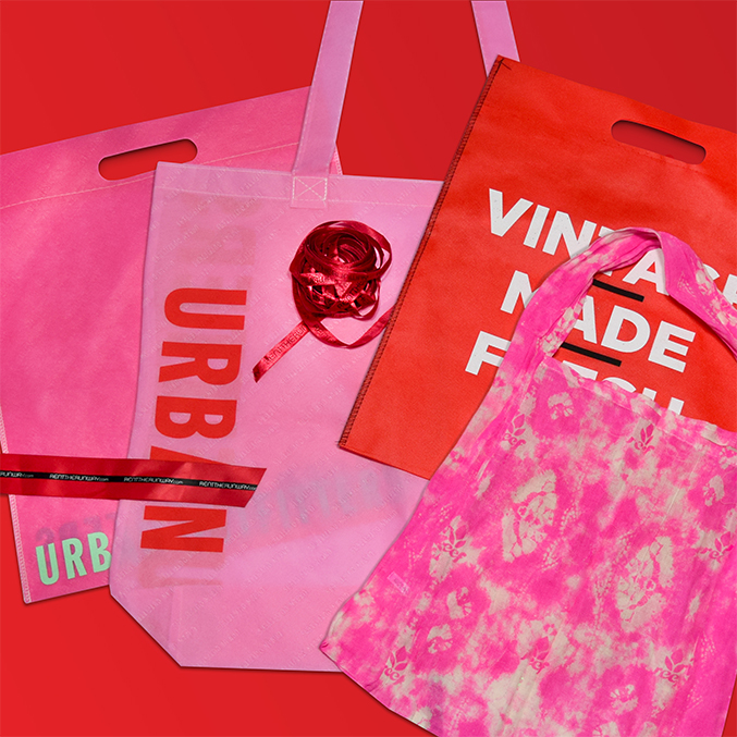 A collection of colorful shopping bags on a red background. The bags feature various designs, including solid colors, text, and tie-dye patterns. A red ribbon is coiled on one of the bags.