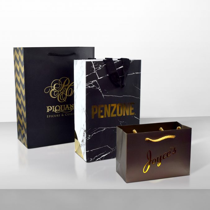 Three elegant shopping bags are displayed on a surface. The bags feature different designs: one with a black and gold pattern, another with a marble look and PENZONE text, and the third in brown with Joyces in gold lettering.