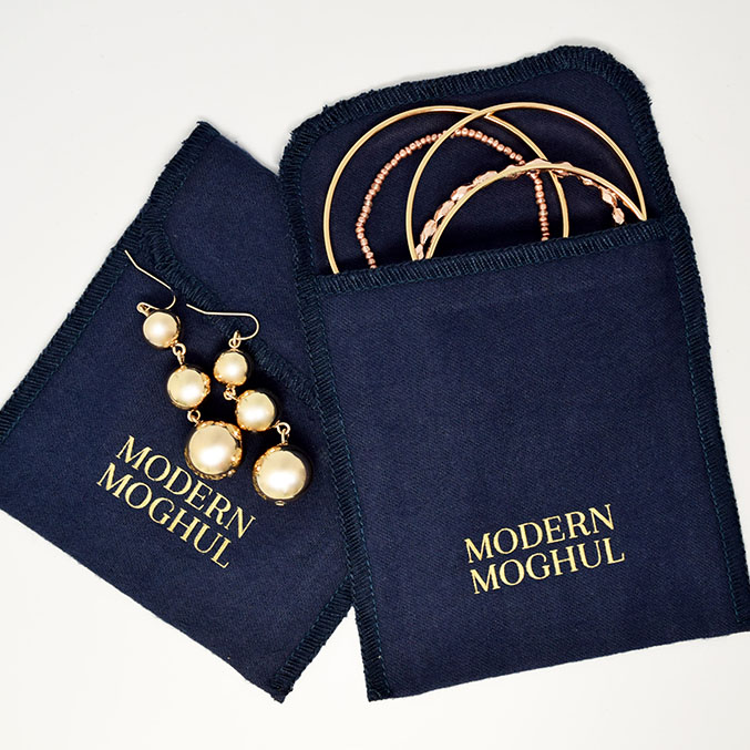 Two navy blue pouches with Modern Moghul text hold three gold bangles and a pair of pearl drop earrings. The pouches are open, revealing the jewelry inside.