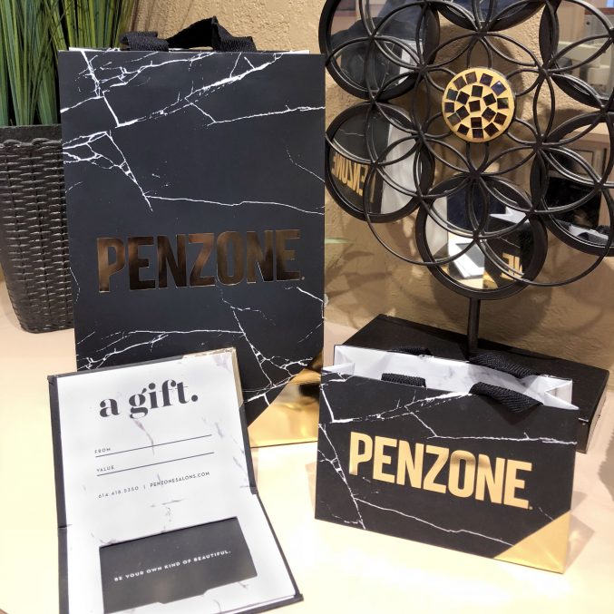Black and gold Penzone gift bags are displayed alongside a decorative sculpture. An open card with a gift is visible, featuring lines for to, from, and value, promoting a gift service. A potted plant is partially visible in the background.
