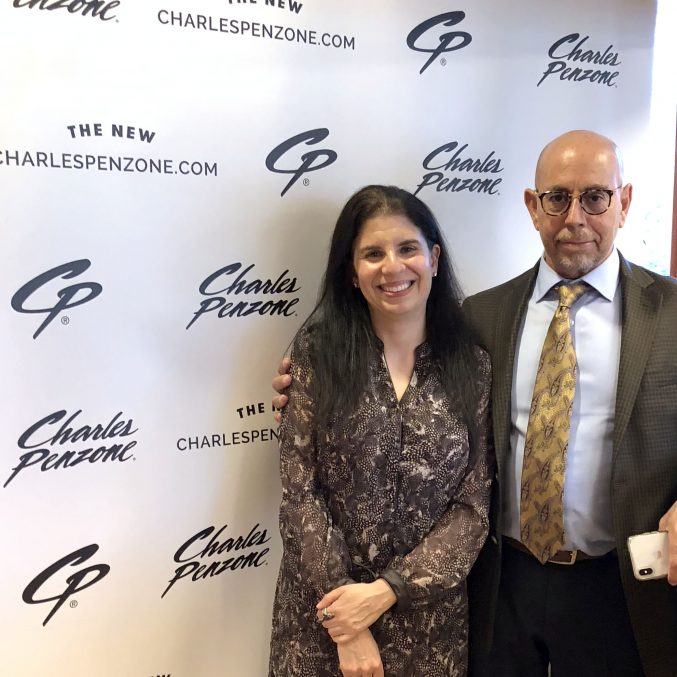 Two people stand smiling in front of a branded backdrop with Charles Penzone and a logo repeated across it. The person on the left is wearing a patterned dress, and the person on the right is in a suit and tie with glasses.