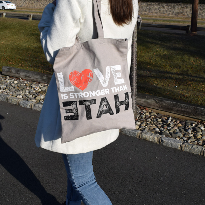 A person wearing a white coat holds a canvas bag over their shoulder. The bag has the text LOVE IS STRONGER THAN with LOVE in red and black letters. The person is standing on a paved path with grass and rocks beside it.