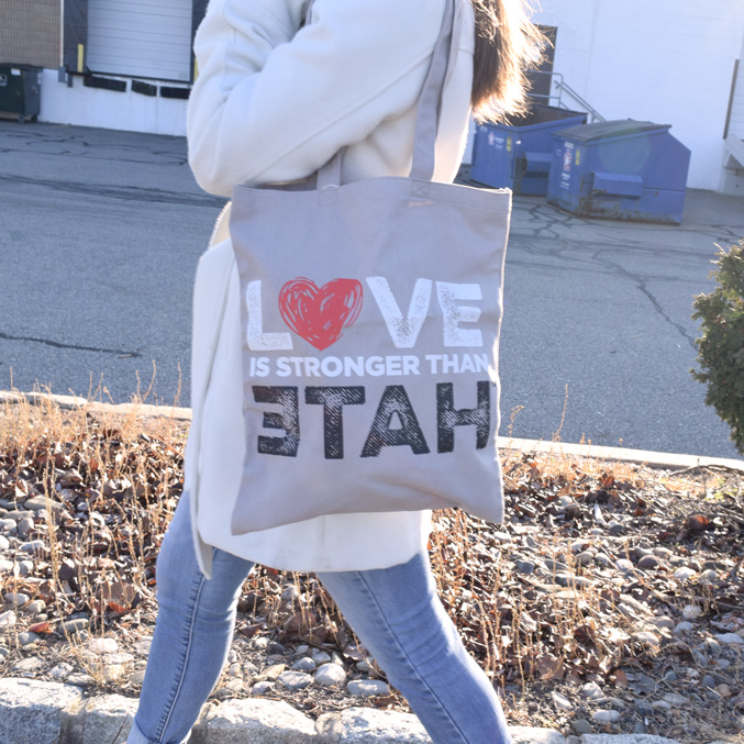 A person walks outside wearing a white coat and jeans, carrying a custom cotton canvas tote bag with the text LOVE IS STRONGER THAN HATE in bold letters, with LOVE partially covered by a red heart design.