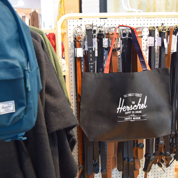A display of various accessories in a shop, featuring a blue backpack on the left and a black tote bag with Herschel Supply Co. branding on the right. Behind them, several belts are hanging on a rack.