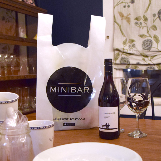 A table set with wine glasses, cups, and a wine bottle labeled Simple Life next to a branded Minibar delivery bag. The room features a dark wall and floral curtains in the background.