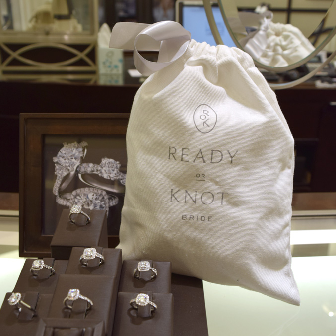 A display of diamond engagement rings on a counter, with a white pouch labeled Ready or Knot Bride beside them. A photograph of two rings is in the background.