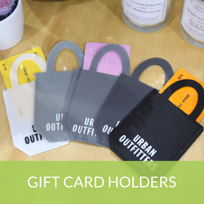 A selection of colorful Urban Outfitters gift card holders on a wooden surface. The holders are grey, black, and white, each containing a colorful card. Above, potted plants and candles are partially visible. Text at the bottom reads GIFT CARD HOLDERS.