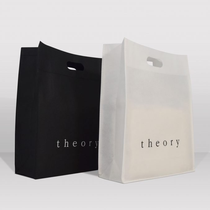 Two shopping bags with cutout handles are displayed side by side. The black bag is on the left and the white bag is on the right. Both bags feature the word theory in lowercase letters. The background is a simple light gray.