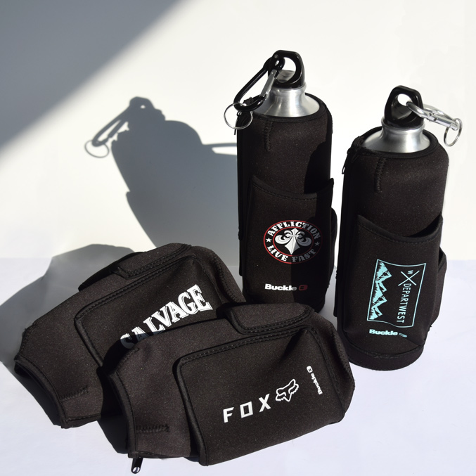 Two insulated water bottles with carabiners, covered in black sleeves featuring various brand logos, are standing upright. Two matching black cloth cases, also with logos, lie in the foreground on a white surface.