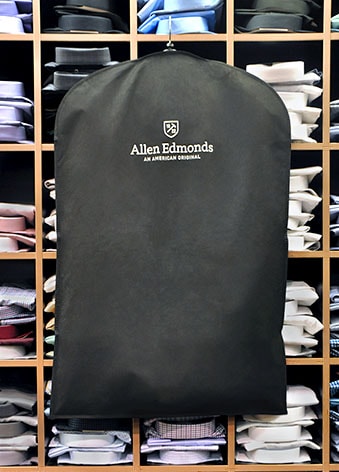 A black custom garment bag branded with Allen Edmonds: An American Original hangs in front of a wall of cubbies filled with folded shirts in various colors and patterns.