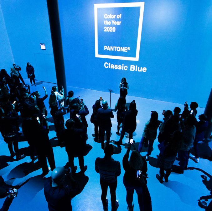 People gather in a room illuminated with blue light, taking photos and listening to a speaker. On the wall is the text Color of the Year 2020 PANTONE Classic Blue.