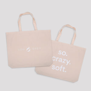 Two beige tote bags on a gray background. One bag displays a logo and the text cool carry, celebrating Earth Day 2020, while the other reads so. crazy. soft. Both feature long handles for easy carrying.