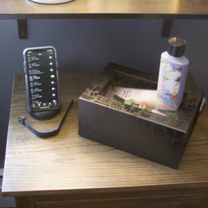A smartphone displaying a schedule rests on a wooden nightstand beside a fitness tracker, a decorative box, and a floral-designed lotion bottle. Above, a shelf provides home storage for various essentials.