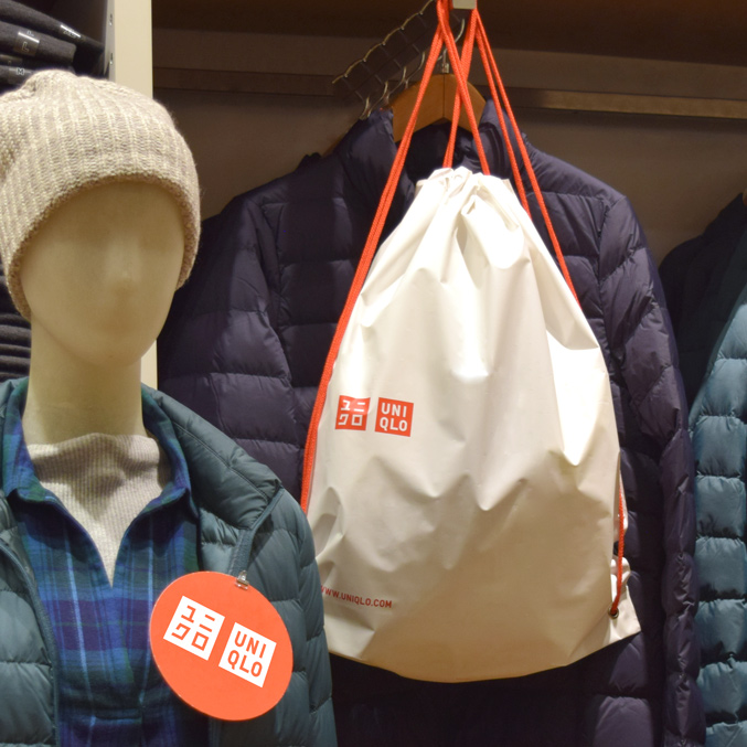 A mannequin wearing a gray beanie and teal plaid shirt under a teal down jacket. A white plastic merchandise bag with handles, featuring red Uniqlo branding, hangs on a hook next to a navy jacket.