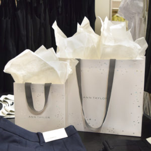 Two Ann Taylor shopping bags, reminiscent of Holiday 2020 cheer, emerge with tissue paper from the tops. The bags rest on a dark surface with neatly folded clothing nearby, while the blurred background showcases more elegant apparel.