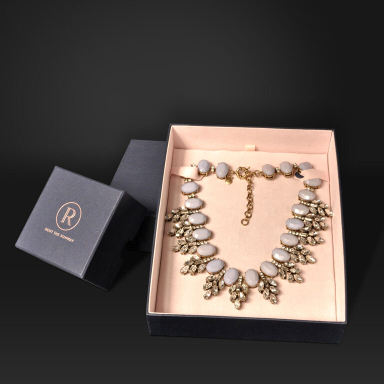 A gold and white statement necklace is elegantly displayed in an open black jewelry box with a beige interior featuring custom necklace box inserts and an R logo on the lid. The necklace showcases oval white stones and dangling gold embellishments, adding a touch of luxury.