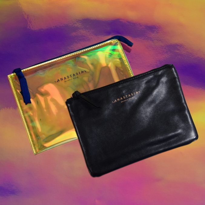 Two cosmetic pouches with zippers, one iridescent and one black, are displayed. Both have JANASTASIA branding. The background is a vibrant blend of pink, orange, and purple hues.