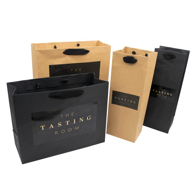Four shopping bags from The Tasting Room, two in black and two in brown. Each bag features black handles and a logo displayed on the front. They are arranged in a neat line, showcasing the brand name.