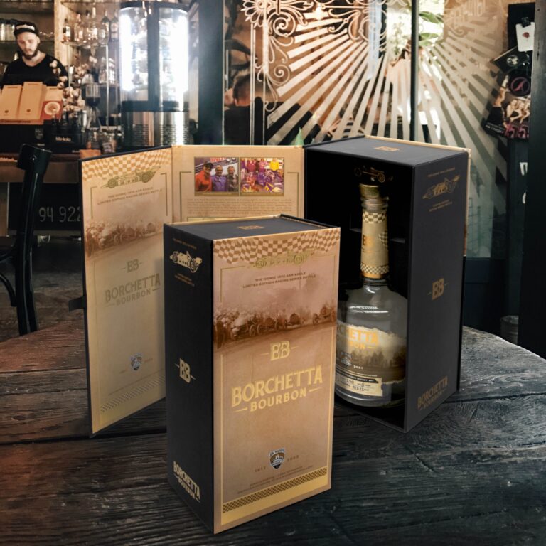 A bottle of Borchetta Bourbon sits elegantly on a wooden table, its custom packaging—a sleek black and gold box—partially enclosing it. The dimly lit bar background adds a touch of sophistication with decorative elements enhancing the scene.