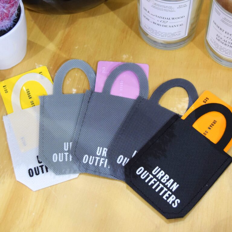 Four small Urban Outfitters gift bags serve as unique gift card holders in black, gray, and white, placed on a wooden surface. Each bag contains a gift card with different values, while candles and a potted plant are partially visible in the background.