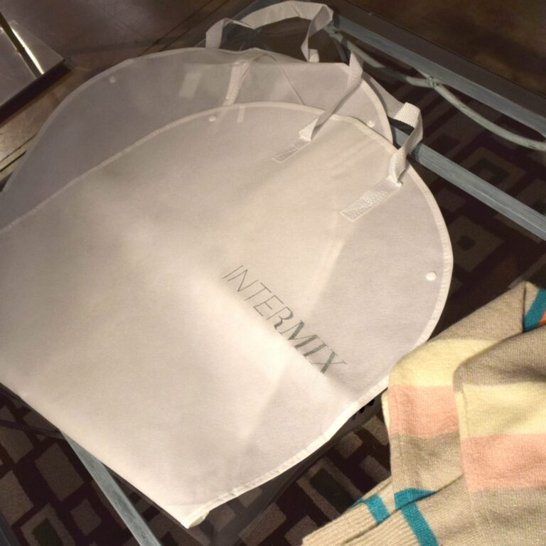 A close-up of two white custom garment bags labeled INTERMIX resting on a glass table. Next to them, a neatly folded striped sweater in pastel colors is partially visible. The geometric patterned surface of the table adds a modern touch to the scene.