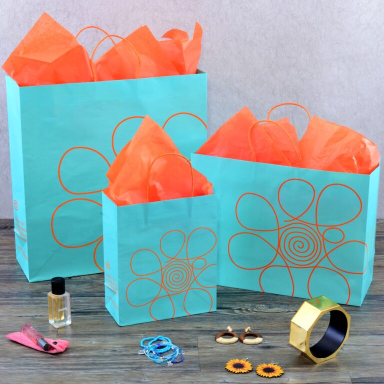 Displayed on a wooden floor, three turquoise custom wholesale shopping bags boast orange floral designs and are filled with vibrant orange tissue paper. In front of them sit a collection of treasures: a gold bracelet, earrings, blue bracelets, nail polish, and pink lip gloss.