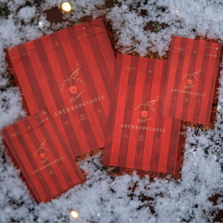 Four red-striped gift bags with the word Anthropologie and Make Merry on them are displayed on a snowy surface. The sizes of the bags vary, creating a festive holiday atmosphere.